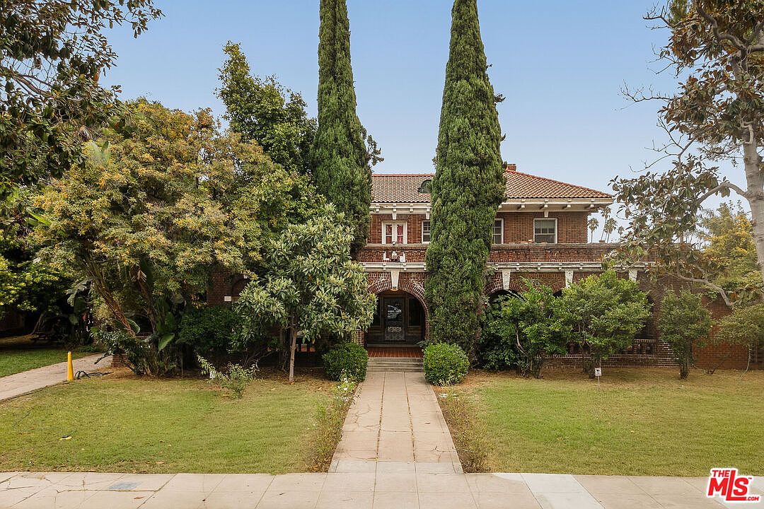 The Rives Mansion, a 1913 Beaux-Arts Mediterranean estate on 1130 Westchester Place in Los Angeles. Two cypress trees flank an arched brick portico at the entry.